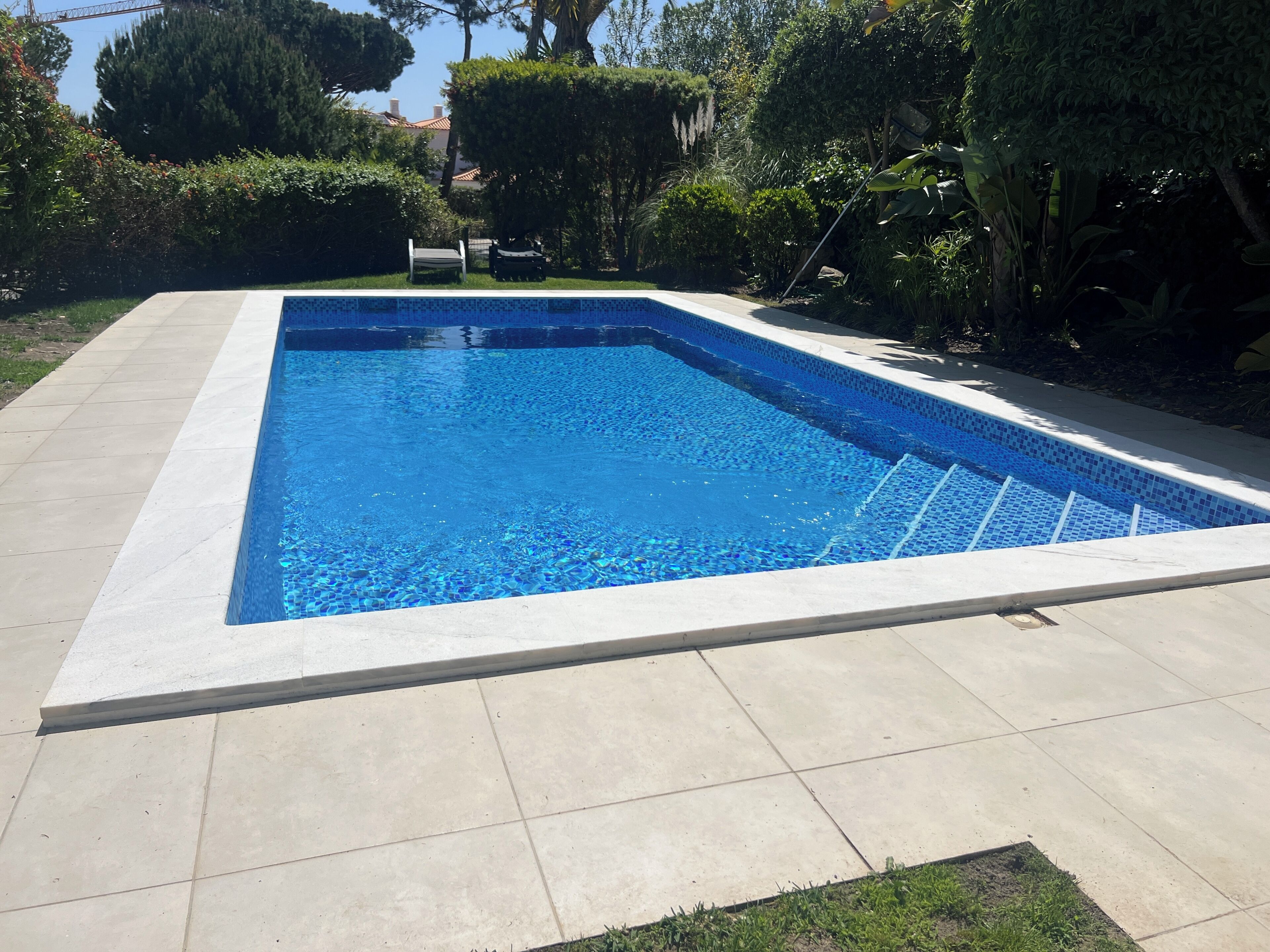 Uma piscina retangular com borda azulejada, rodeada por um pátio pavimentado e vegetação variada.