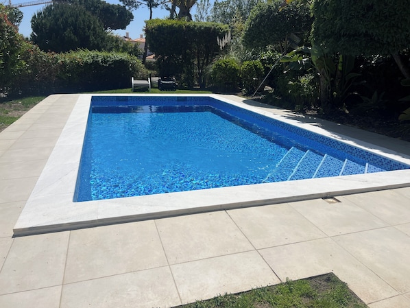 A rectangular swimming pool with a tiled edge, surrounded by a paved patio and various greenery.