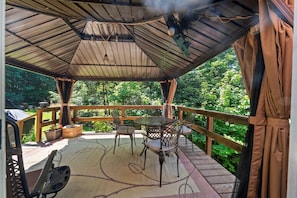 A covered outdoor deck with a table and chairs, surrounded by a wooden railing and lush greenery. - Riverfront Log Home - 28 Acres - Fire Pit (Warner)