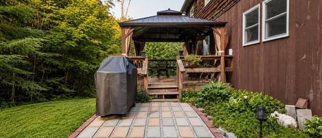 Una terraza de madera con un área de estar techada, una parrilla y un camino que lleva a una casa de revestimiento de madera.