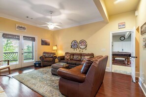 Ein Wohnzimmer mit einem braunen Ledersofa, zwei Sesseln, einem Couchtisch und einem gemusterten Teppich. Eine Deckenlampe, eine Wanduhr und ein Schild mit der Aufschrift 'CAUTION MAN CAVE' sind ebenfalls vorhanden.