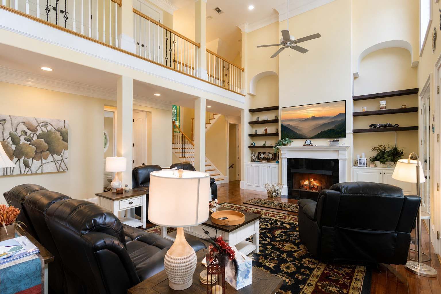 A spacious living room with a fireplace, leather sofas, a high ceiling, and a staircase leading to an upper level.