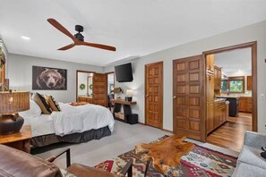 Une chambre à coucher comprenant un lit, un ventilateur de plafond, une télévision et une porte en bois donnant sur une salle de bain.