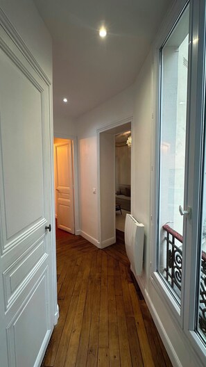 Ein Korridor mit Holzboden, weißen Wänden und einer Tür zu einem Raum mit Heizkörper und Fenster mit Metallgitter.