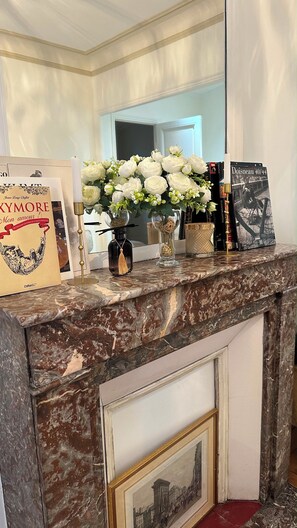 Ein Kamin mit Marmorverkleidung, darauf ein gerahmtes Bild, eine Vase mit weißen Blumen und ein Buch mit dem Titel 'Dossier 40/44'.