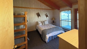 A bedroom with a bunk bed, a bed with a grey and white quilt, a nightstand with a lamp, and a window with a view of the outdoors. - Rio Grande River and Ridge Lodge (South Fork)