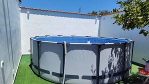 Une piscine ronde, installée à l’extérieur, avec un revêtement intérieur bleu, entourée d’un gazon artificiel vert, adossée à un mur blanc surmonté d’un toit recouvert de tuiles rouges.