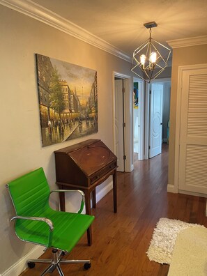 Un couloir doté d’un plancher en bois, d’une chaise verte, d’une table en bois, d’un tableau représentant une scène de rue et d’un lustre moderne.