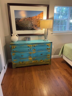 Een kamer met een houten vloer, een turquoise dressoir met meerdere lades, een witte kan, een lamp en een lijst met een landschapsafbeelding.
