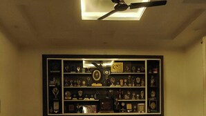 Una sala de estar con ventilador de techo, una mesa de centro de vidrio, dos sofás y una estantería con trofeos en la pared.