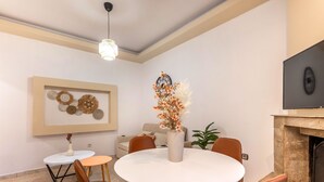 Een eethoek met een ronde tafel, stoelen, een vaas met bloemen, een hangklok en een open haard.