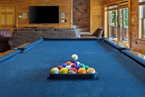 A pool table with balls arranged in a triangle formation, a television mounted on the wall, and a wooden cabin interior with a window.
