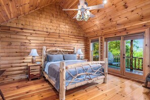 A log cabin bedroom with a wooden bed, two bedside tables, a chandelier, and a view of the outdoors.