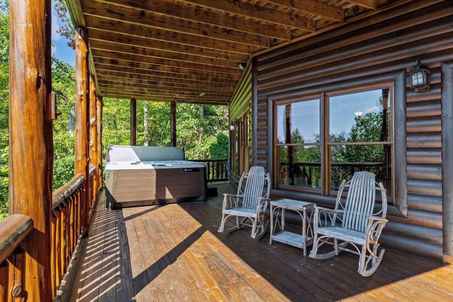 A wooden deck with a hot tub, two rocking chairs, and a large window overlooking a forest.