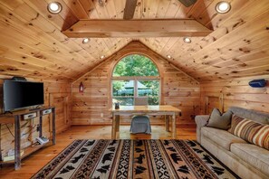 A cozy cabin interior with a wooden ceiling, a television on a wooden stand, a table, a sofa, and a large window with a view of greenery. - Luxury Log Cabin | Epic Views, Hot Tub, Sleeps 12 (Mineral Bluff)