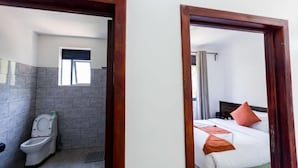 Een badkamer met toilet en douche, een bed met kussens, en een raam met gordijnen.