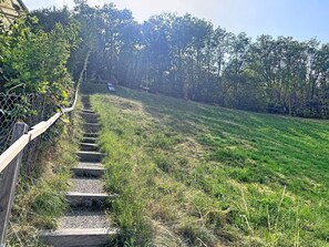 Eine Holztretter führt einen grasbewachsenen Hang hinauf, umgeben von einem Drahtzaun und dichter Vegetation.