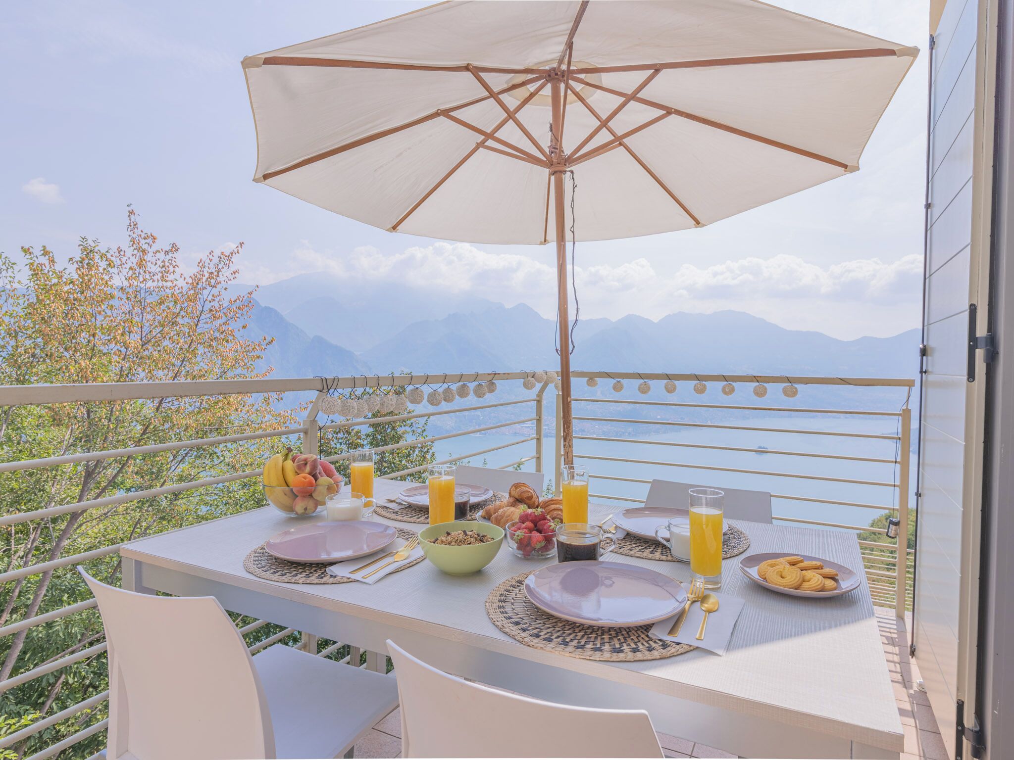 Una tavola imbandita per la colazione con vista sulle montagne, un ombrellone bianco e una ringhiera.