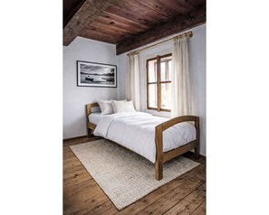 Una cama con estructura de madera, ropa de cama blanca y una alfombra sobre piso de madera.