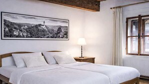 A bedroom with a large bed, a bedside lamp, a window with curtains, and a framed landscape painting on the wall.