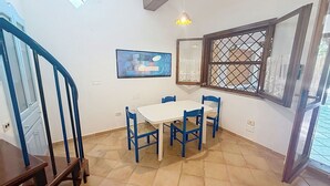 Ein Essbereich mit einem weißen Tisch und blauen Stühlen, ein Fenster mit Gittermuster sowie eine Treppe mit blauen Geländern.