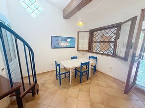 Ein Essbereich mit einem weißen Tisch und blauen Stühlen, ein Fenster mit Gittermuster sowie eine Treppe mit blauen Geländern.