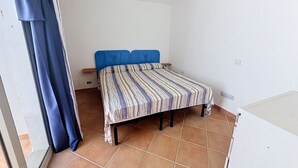 Ein Schlafzimmer mit einem Bett, einem Fenster mit Vorhängen, einer weißen Wand und einem gefliesten Boden.