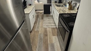 Cocina con refrigerador de acero inoxidable, estufa y microondas. Hay una pequeña isla con un fregadero y una silla. El piso es de madera, y hay un tapete que lleva a una puerta.