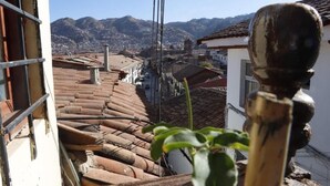 Vista desde una ventana sobre una calle con tejados de terracota, una planta en primer plano y un paisaje montañoso al fondo.