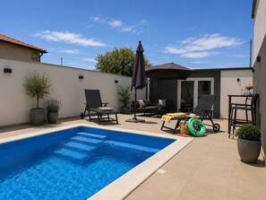 Une piscine entourée de chaises longues et de parasols, le tout bordé d’un mur blanc et accessible par une porte qui donne sur un patio couvert.