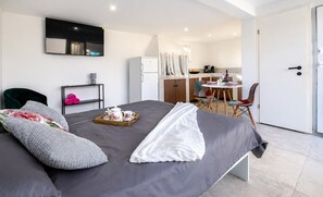 Une chambre d’hôtel moderne avec un grand lit, une petite table sur laquelle est disposé un service à thé, une chaise et, au fond, une kitchenette.