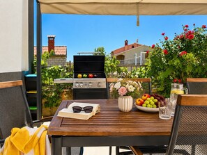 Eine Terrasse im Freien mit Grill, Tisch, Buch, Gläsern, Blumenvase und Obstschale.