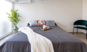 Ein ordentlich bezogenes Bett mit grauer Bettdecke, einer weißen Zudecke und einem Tablett mit Teekanne und Tassen. Ein grüner Sessel und ein Blumentopf stehen am Fenster.