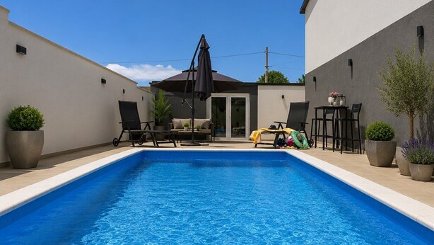 Ein rechteckiger Swimmingpool mit klarem, blauem Wasser, umgeben von einer weißen und einer grauen Mauer. Außerhalb des Poolbereichs stehen Liegestühle und ein Tisch mit Stühlen.