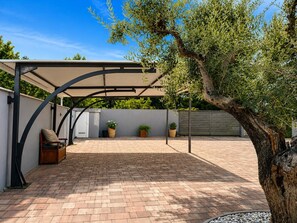 Ein überdachter Patio-Bereich mit Terrazzo-Boden, einer Sitzgelegenheit mit Kissen und Topfpflanzen.