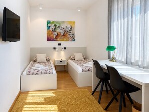 Ein kleines Zimmer mit zwei Einzelbetten, einem Schreibtisch mit Stuhl, einem an der Wand befestigten Fernseher und einem farbenfrohen, abstrakten Gemälde über den Betten.