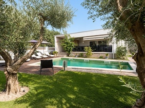A modern outdoor pool area with a lounge chair, a small table, and a building with large windows. - Holiday House Pool Retreat (Pomer)