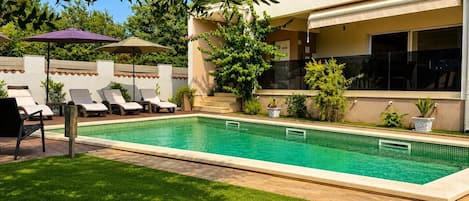 A swimming pool with lounge chairs and a covered area, surrounded by greenery and a building.