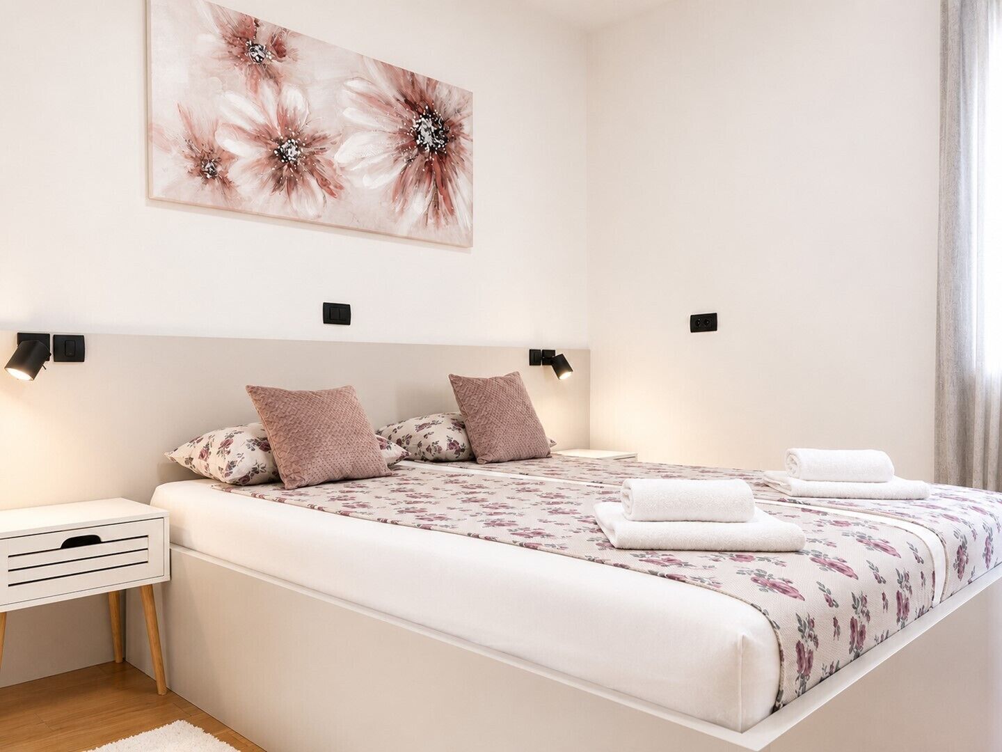 A bedroom with a bed, pillows, a nightstand, a wall-mounted light, and a window.