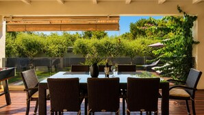 Ein Essbereich im Freien mit einem Glastisch und Korbstühlen, mit Blick auf einen Garten mit Bäumen und einer Pergola.