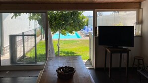 A room with a wooden ceiling, a TV on a stand, a table with a bowl, and a view of a backyard with a tree and a swimming pool. - Vivenda Ribeiro: House with Mountain View, Private Pool and Wi-Fi (Curvos)