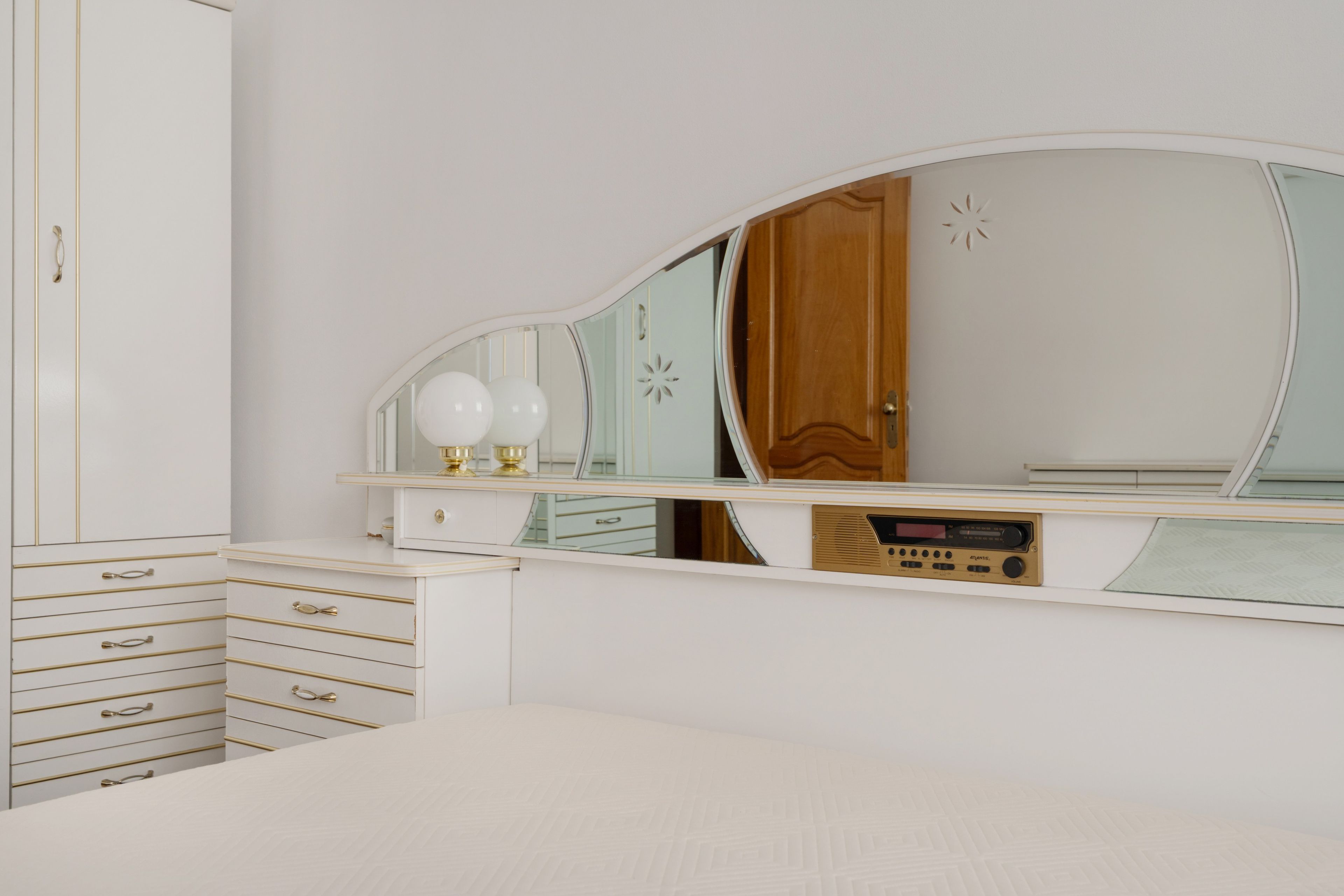 Un dormitorio con una cama, un aparador con cajones, una tocador con espejo y una radio.