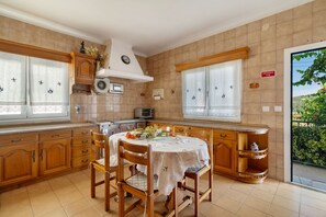 A kitchen with wooden cabinets, a dining table with chairs, a built-in oven, and a window with an emergency exit sign. - Vivenda Ribeiro: House with Mountain View, Private Pool and Wi-Fi (Curvos)