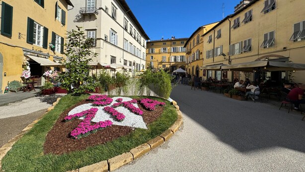 Calle con edificios, mesas al aire libre y un cantero de flores.