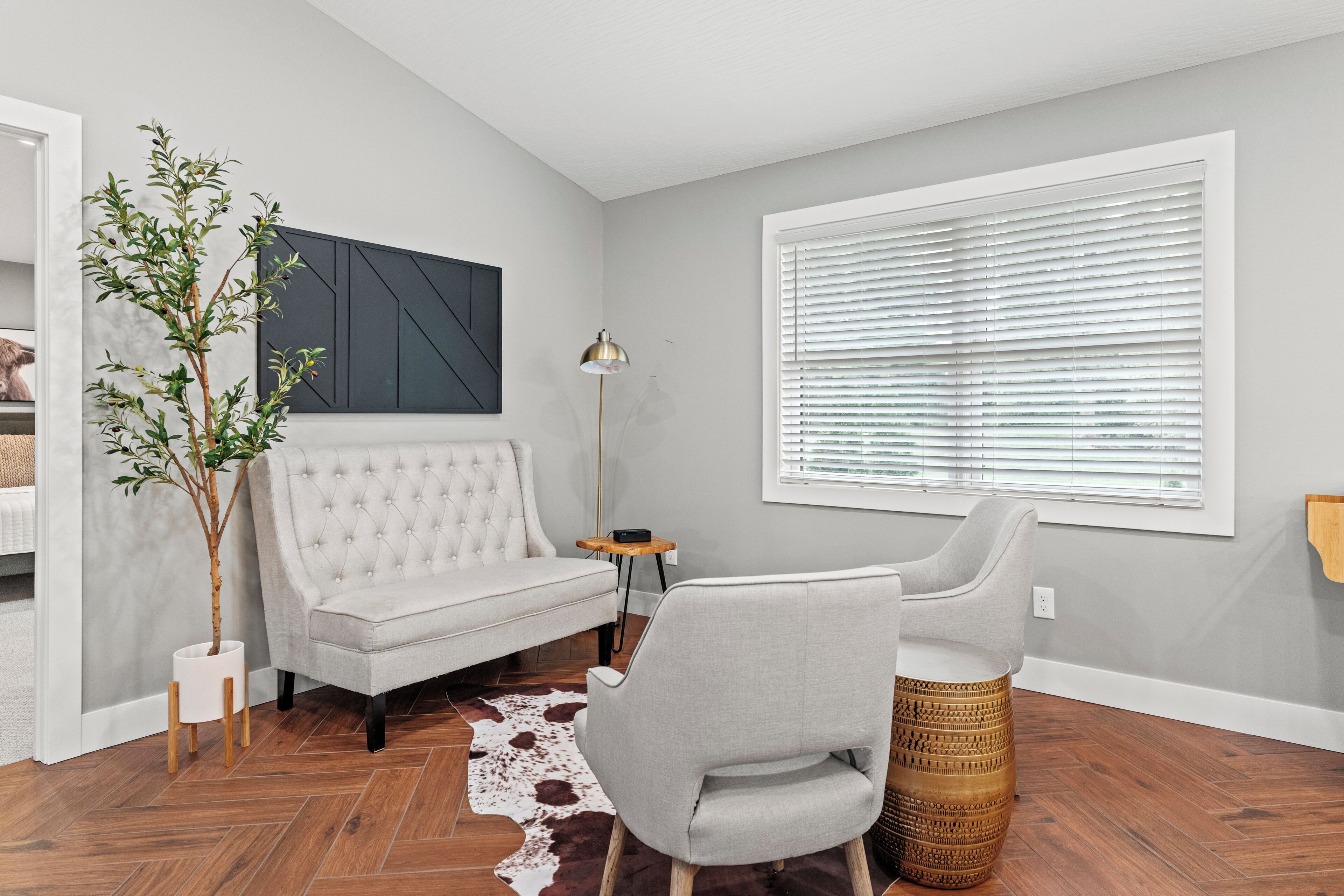 A modern living room with a tufted sofa, a side table, a lamp, a large window with blinds, a cowhide rug, and a potted plant.