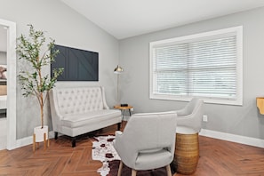 A modern living room with a tufted sofa, a side table, a lamp, a large window with blinds, a cowhide rug, and a potted plant.