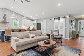 A modern living room with a sofa, a patterned armchair, a coffee table, and a dining area with chairs and a kitchen in the background.