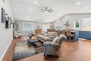 A spacious living area with a sofa, coffee table, and patterned armchair. A kitchen area is visible with a dining table and bar seating.
