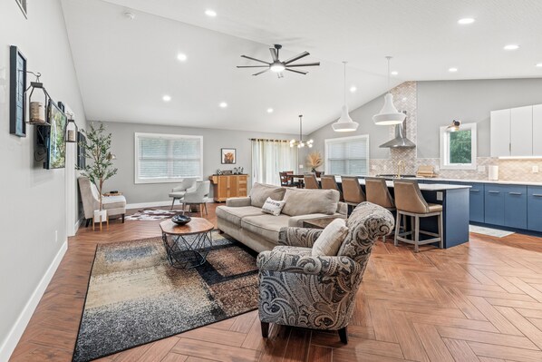 A spacious living area with a sofa, coffee table, and patterned armchair. A kitchen area is visible with a dining table and bar seating.