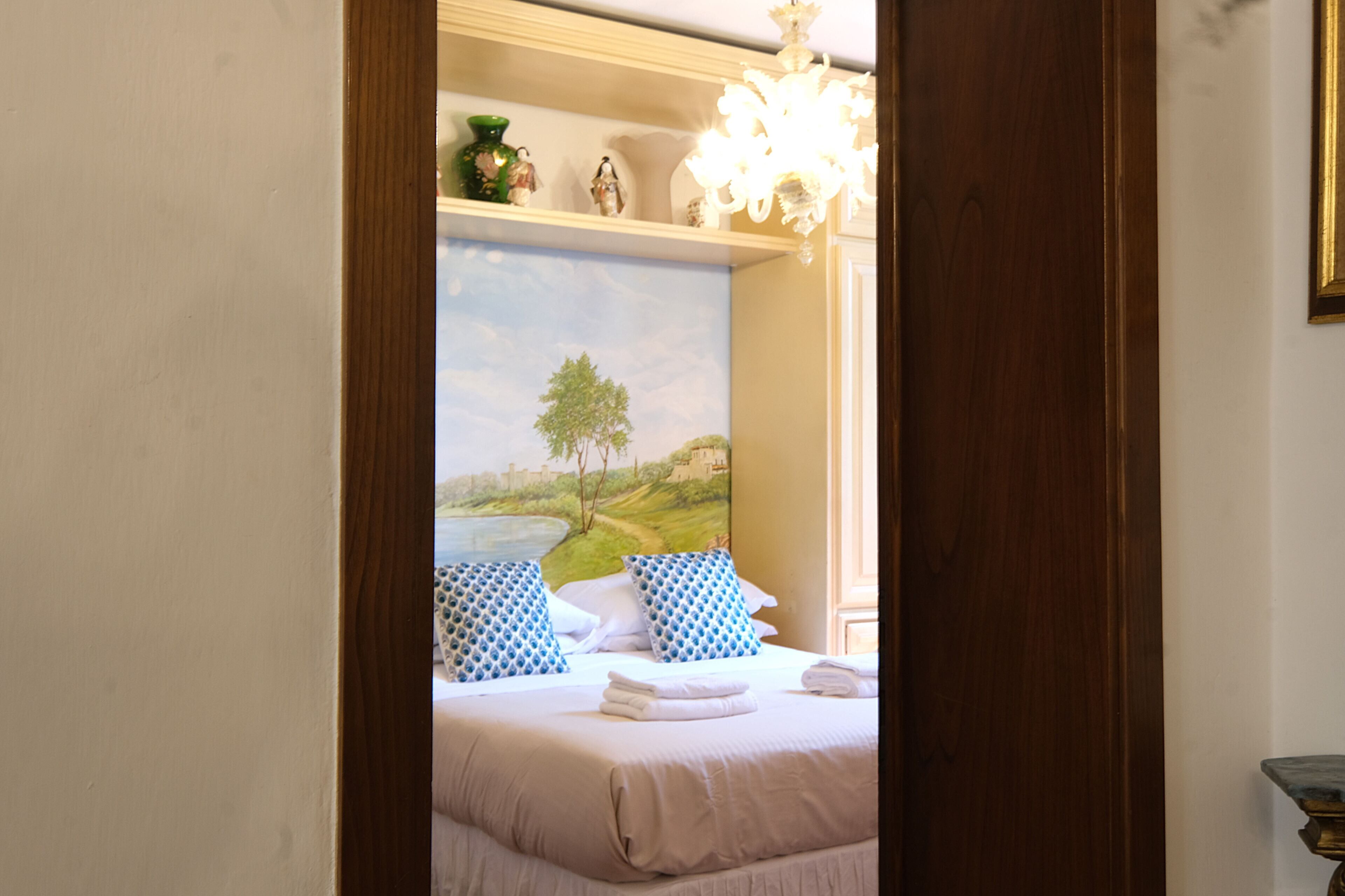 Quarto com cama, travesseiros, pintura de paisagem e lustre.